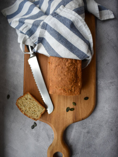 Drewniana deska na szarym tle. Na niej świeży bochenek chleba zawinięty w biało niebieską ścierkę. Obok nóż i ukrojona kromka.