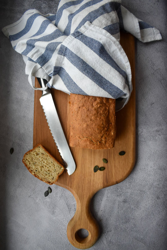 Drewniana deska na szarym tle. Na niej świeży bochenek chleba zawinięty w biało niebieską ścierkę. Obok nóż i ukrojona kromka.