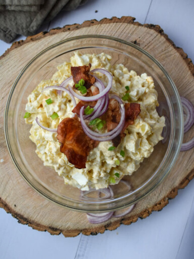 Szklana miska na grubym plastrze drewna, a w niej sałatka ziemniaczana podana z bekonem i czerwoną cebulą