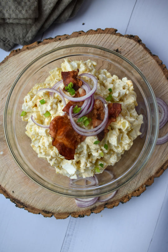Szklana miska na grubym plastrze drewna, a w niej sałatka ziemniaczana podana z bekonem i czerwoną cebulą